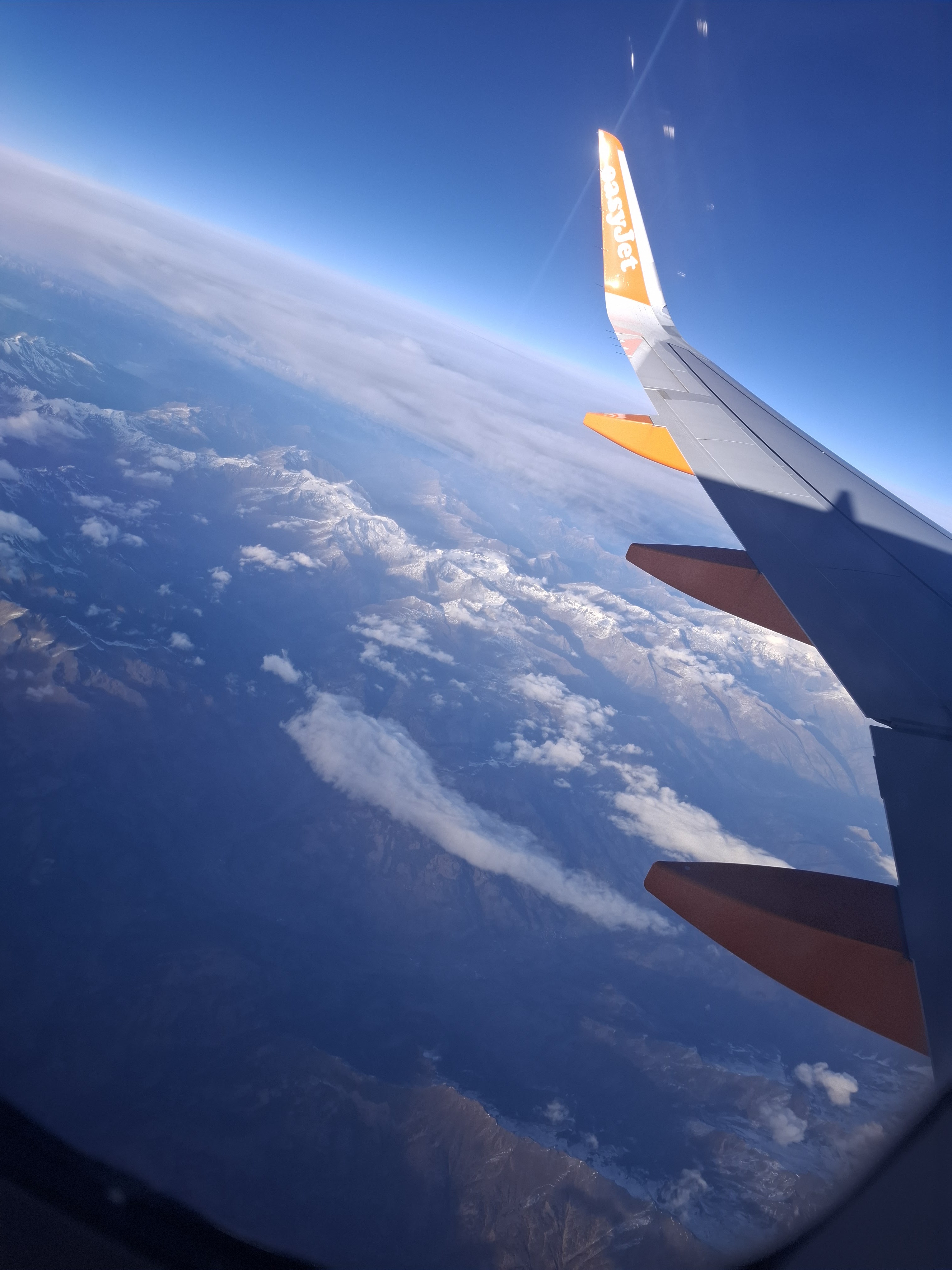 Aerial view from an airplane window showing mountain ranges and valleys under clear skies, with the wing of an easyJet aircraft partially visible.