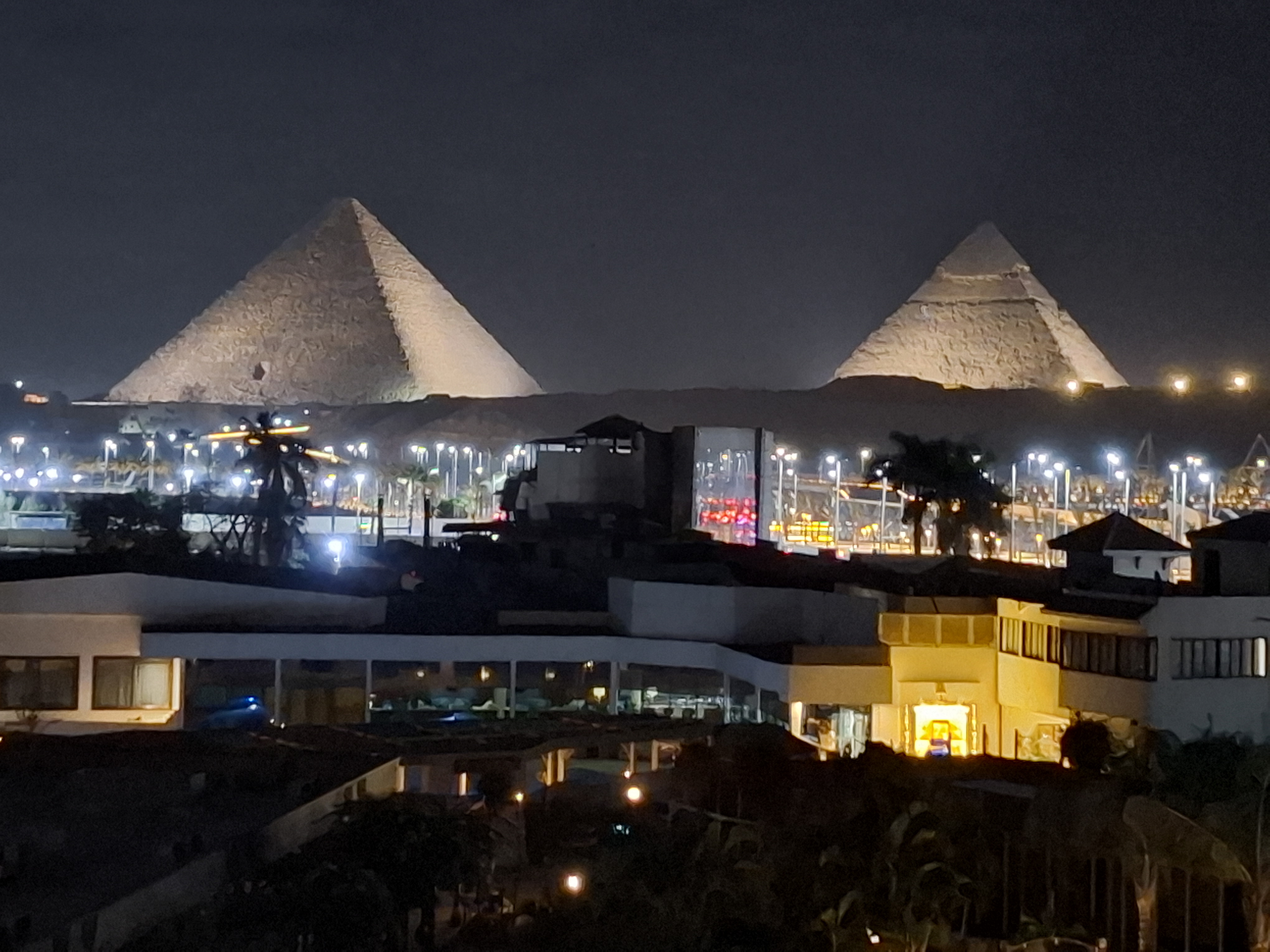 View of the Pyramids of Giza illuminated at night, with surrounding buildings and city lights visible in the foreground.