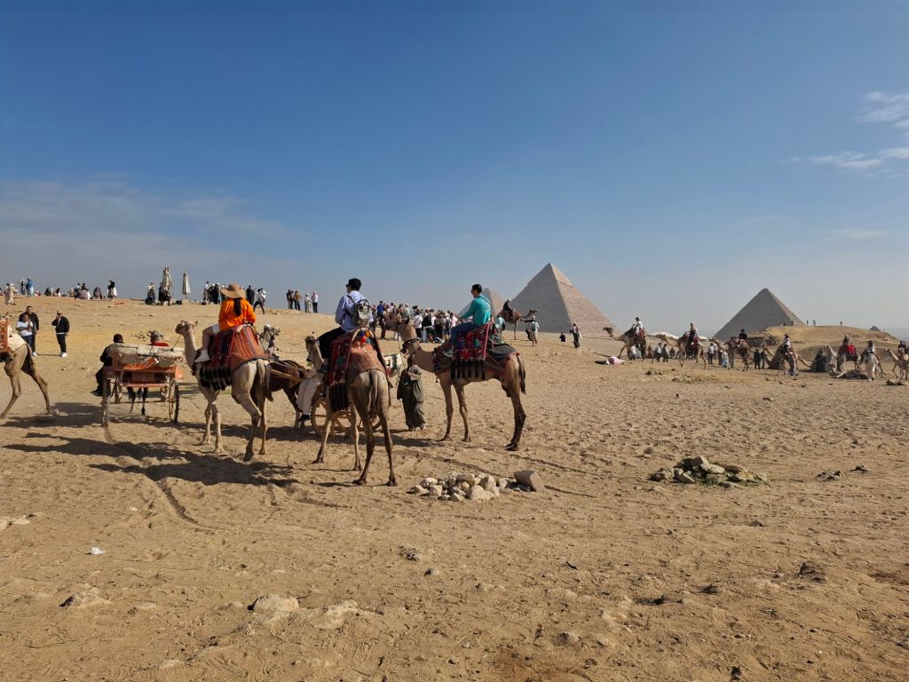 A busy scene at the Pyramids of Giza, showing tourists riding camels and exploring the sandy landscape with the pyramids in the background.