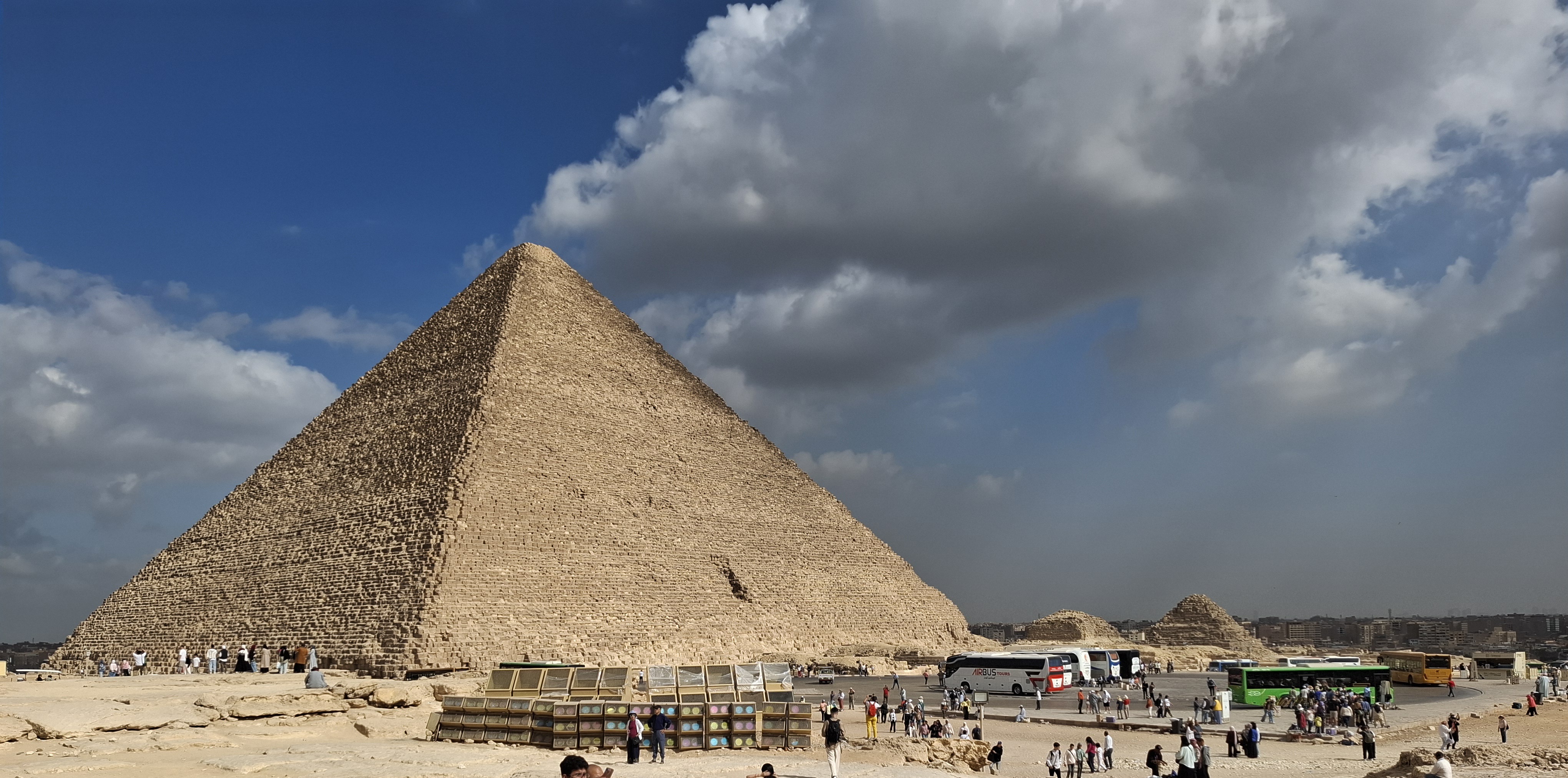 The Great Pyramid of Giza towering against a blue sky with clouds, surrounded by tourists and buses, showcasing the iconic ancient structure and the bustling atmosphere of the Giza plateau.