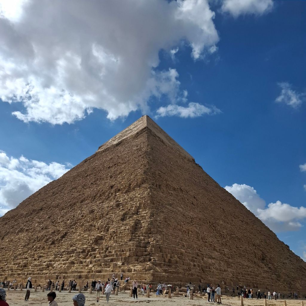 A view of the Great Pyramid of Giza, showcasing its massive limestone structure against a blue sky with fluffy clouds. Tourists are seen exploring the area around the pyramid.