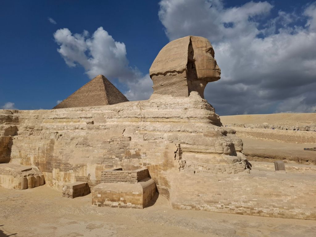 A view of the Great Sphinx with the Pyramid of Khufu in the background, under a partly cloudy sky.