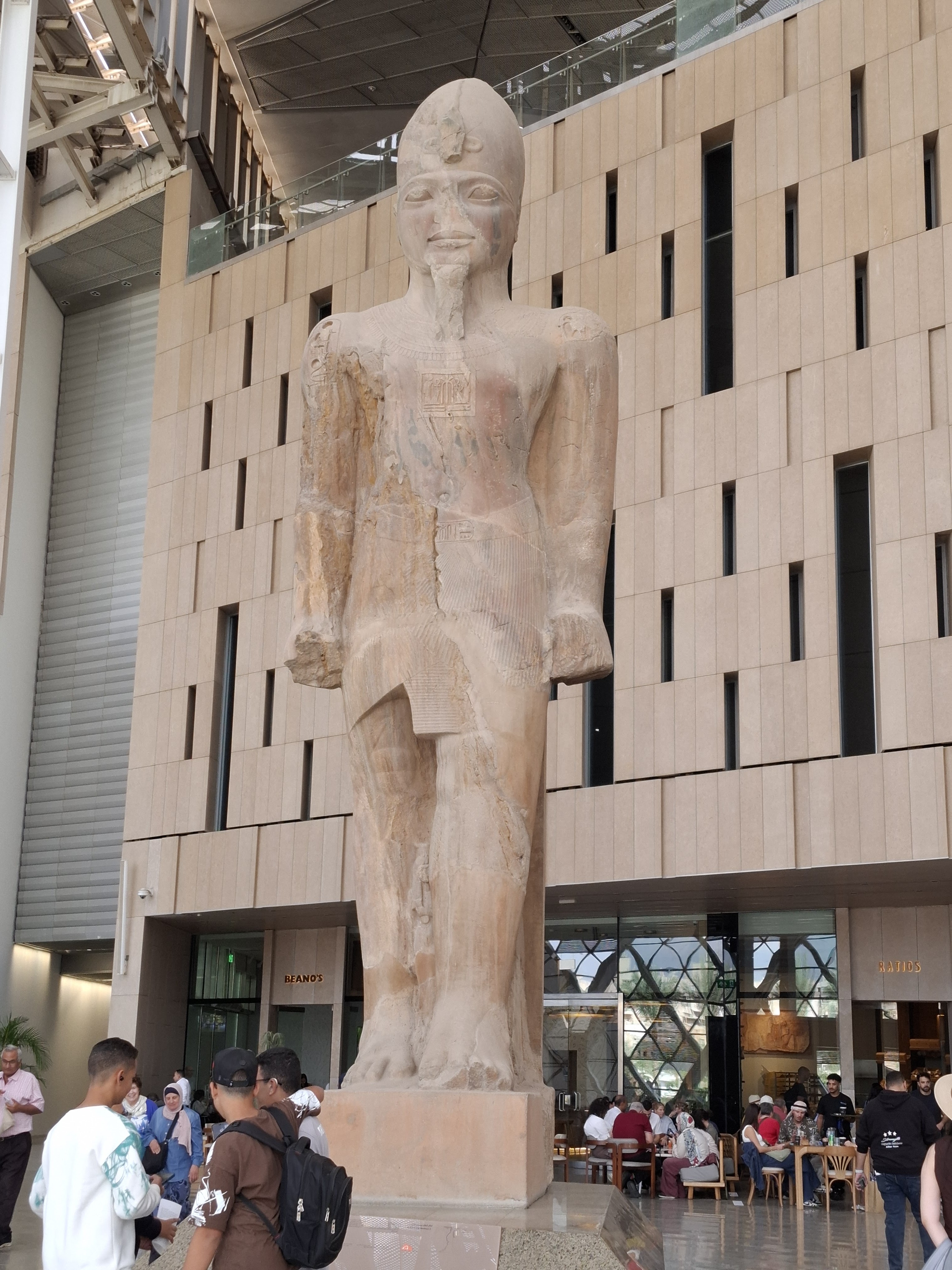 A large ancient statue of Ramses II inside the Grand Egyptian Museum, surrounded by visitors and modern architectural elements.