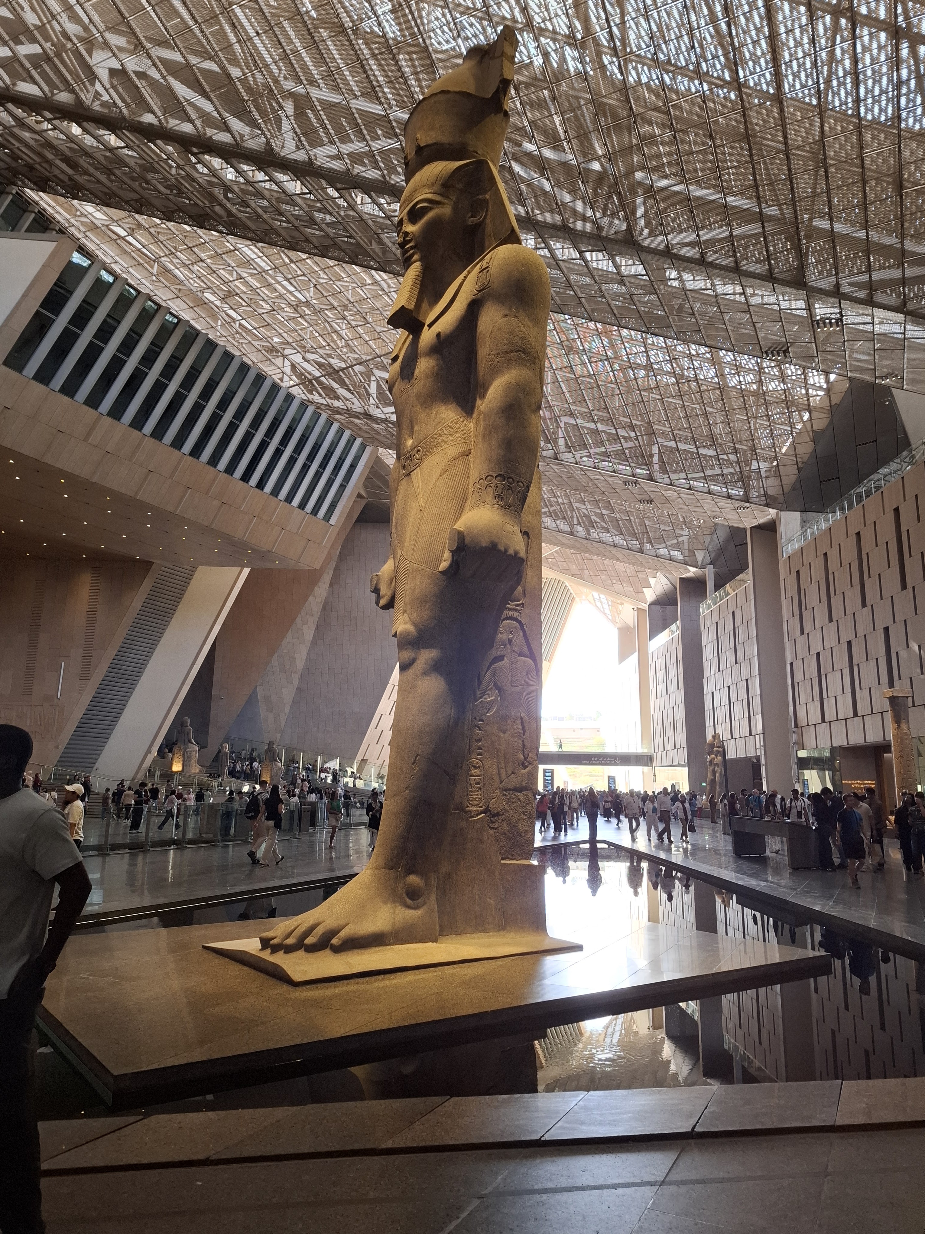 A towering statue of Ramses II stands inside the Grand Egyptian Museum, showcasing ancient Egyptian artistry. The statue is situated in an expansive hall with modern architectural features and a reflective shallow pool in front of it, while visitors can be seen exploring the museum in the background.