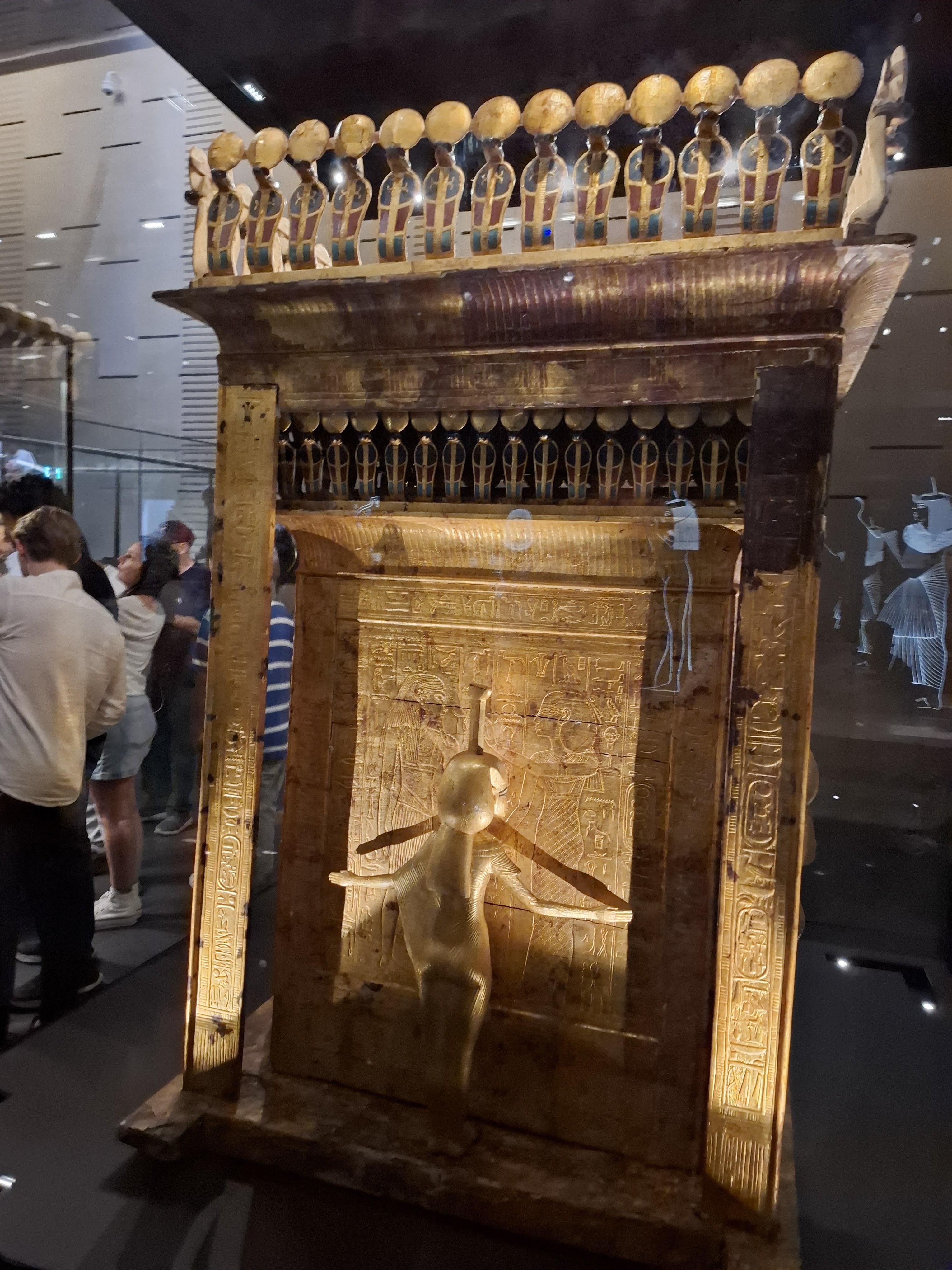 A detailed view of an ancient Egyptian artifact displayed in a museum, featuring intricate carvings and gold elements, surrounded by visitors.