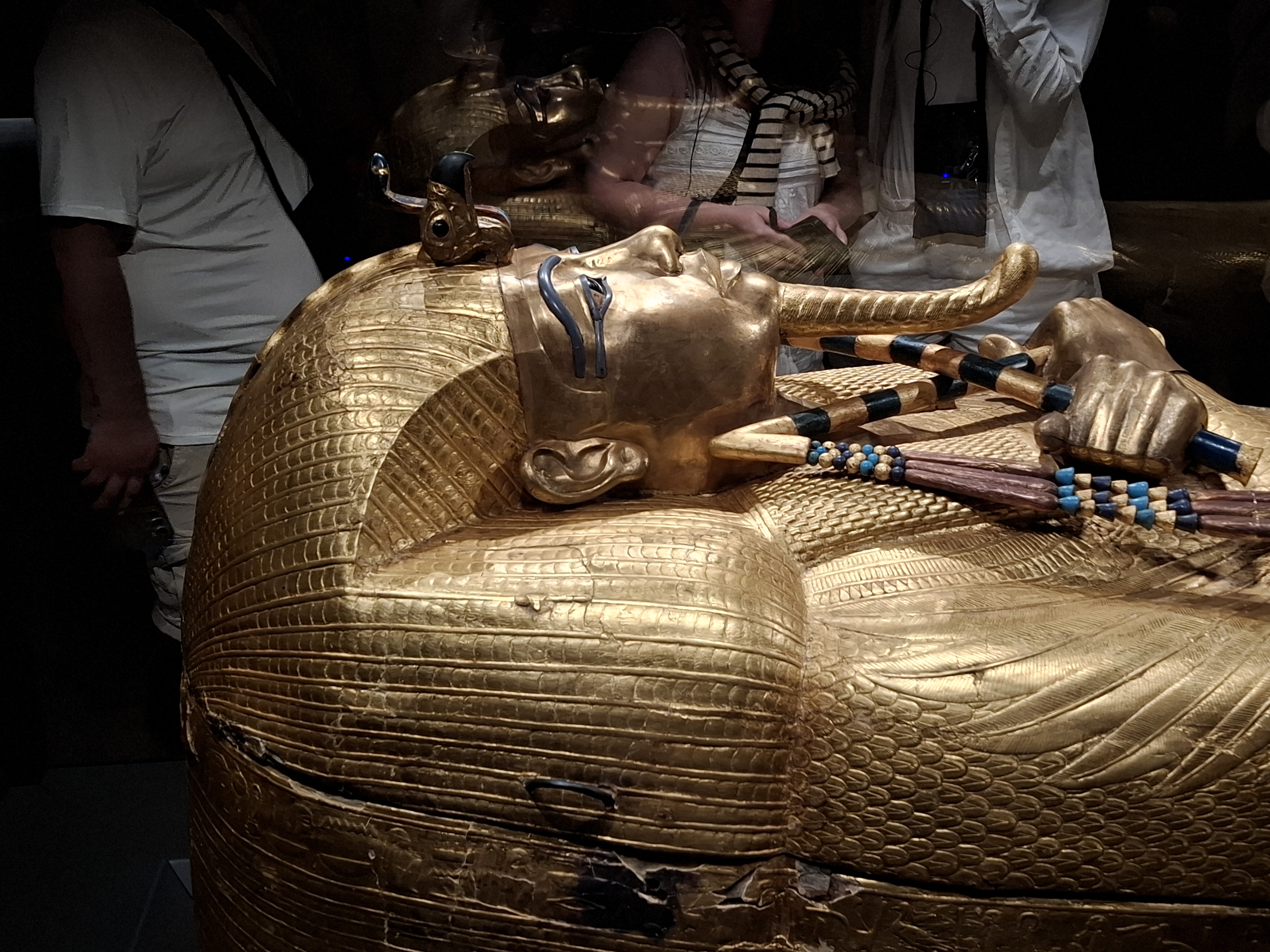 A close-up view of the golden sarcophagus of Tutankhamen, displayed in a museum, showcasing intricate details and decorations, with visitors in the background.