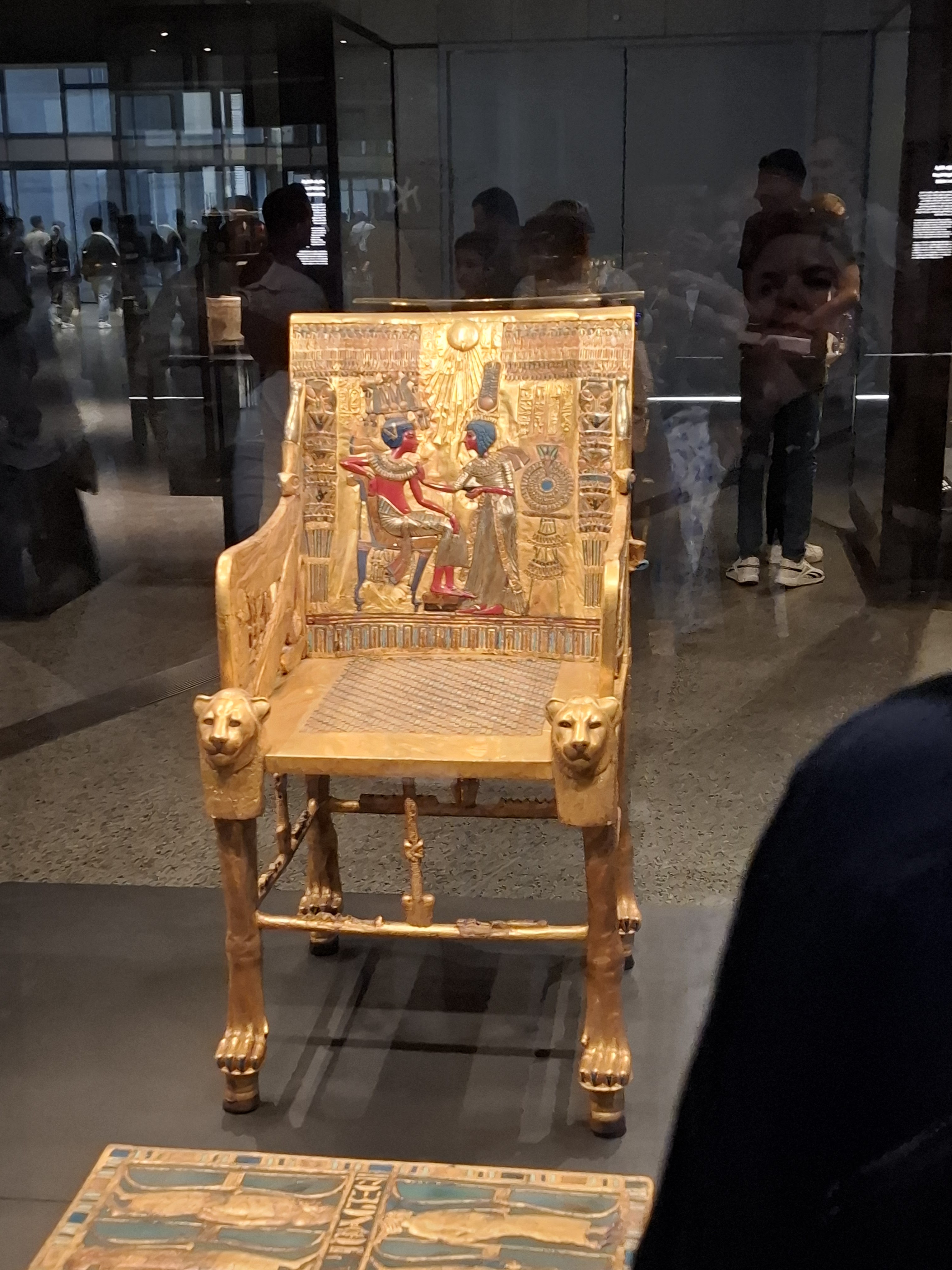 A golden throne in the Grand Egyptian Museum, featuring intricate engravings depicting figures and adorned with lion heads. Reflections of visitors can be seen on the glass display case.