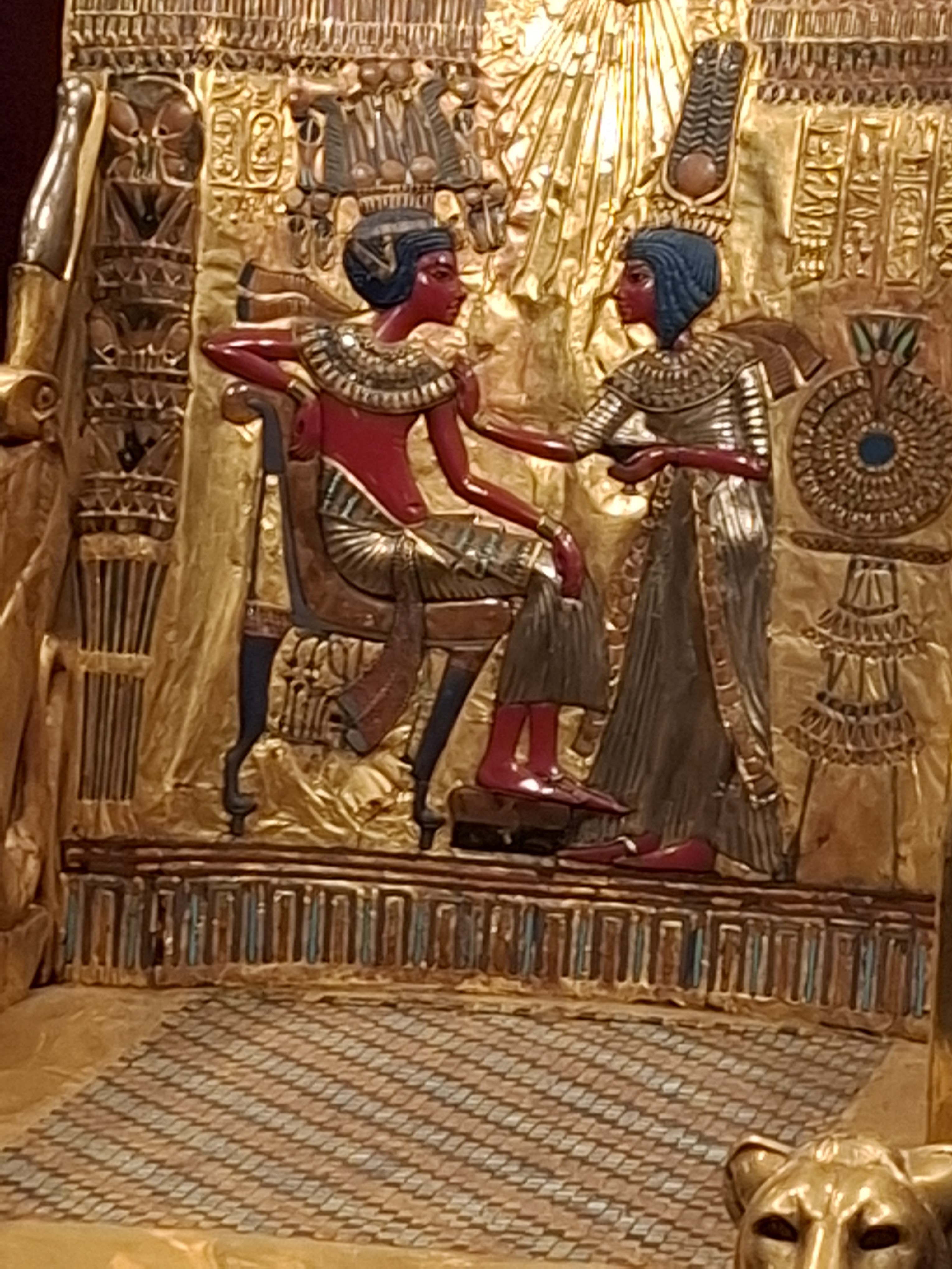 A detailed gold and colored relief depicting two ancient Egyptian figures, one seated and another standing, with intricate ornamental designs in the background.
