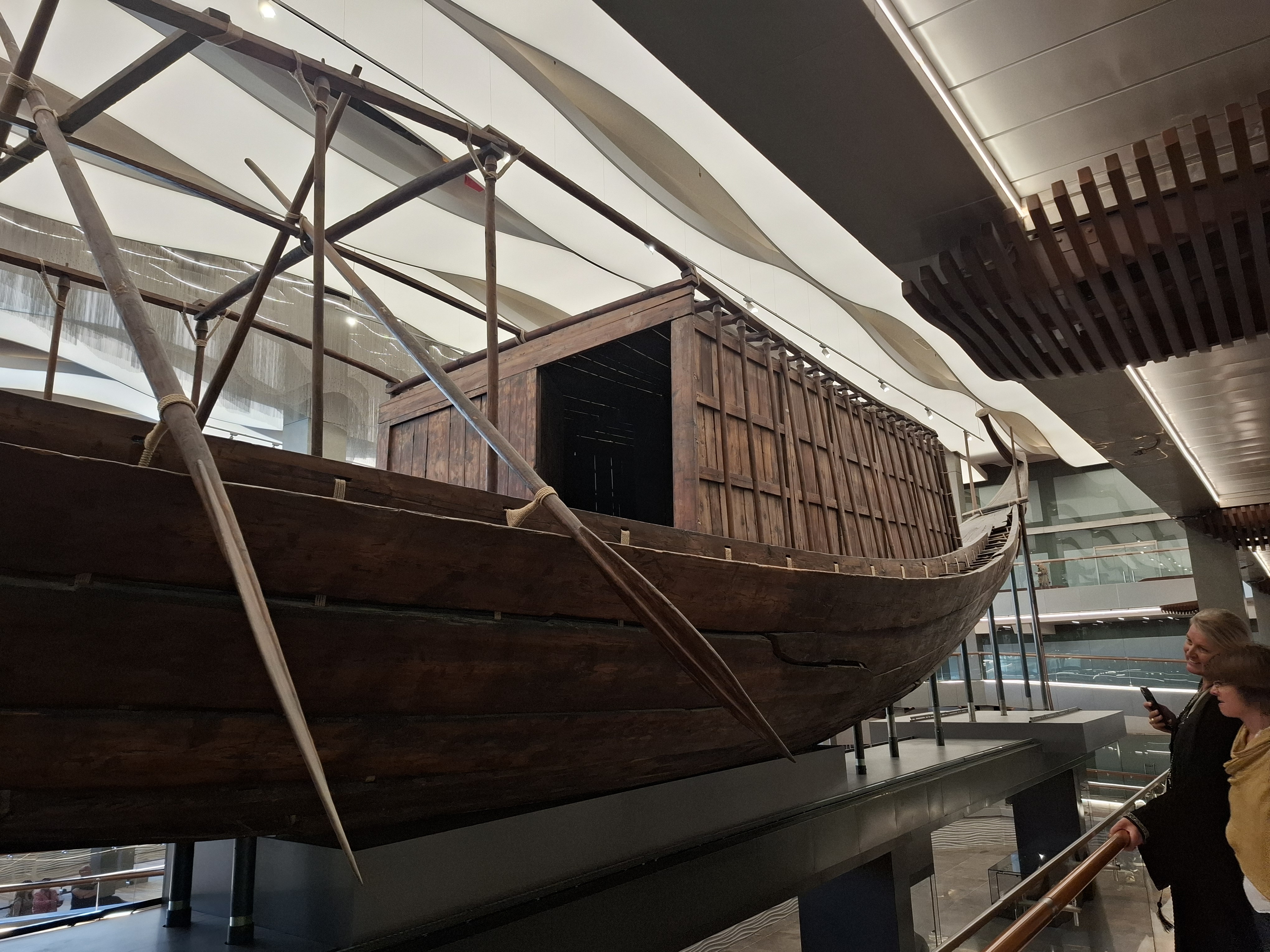 A wooden ancient Egyptian solar boat displayed in a museum, with visitors admiring it.