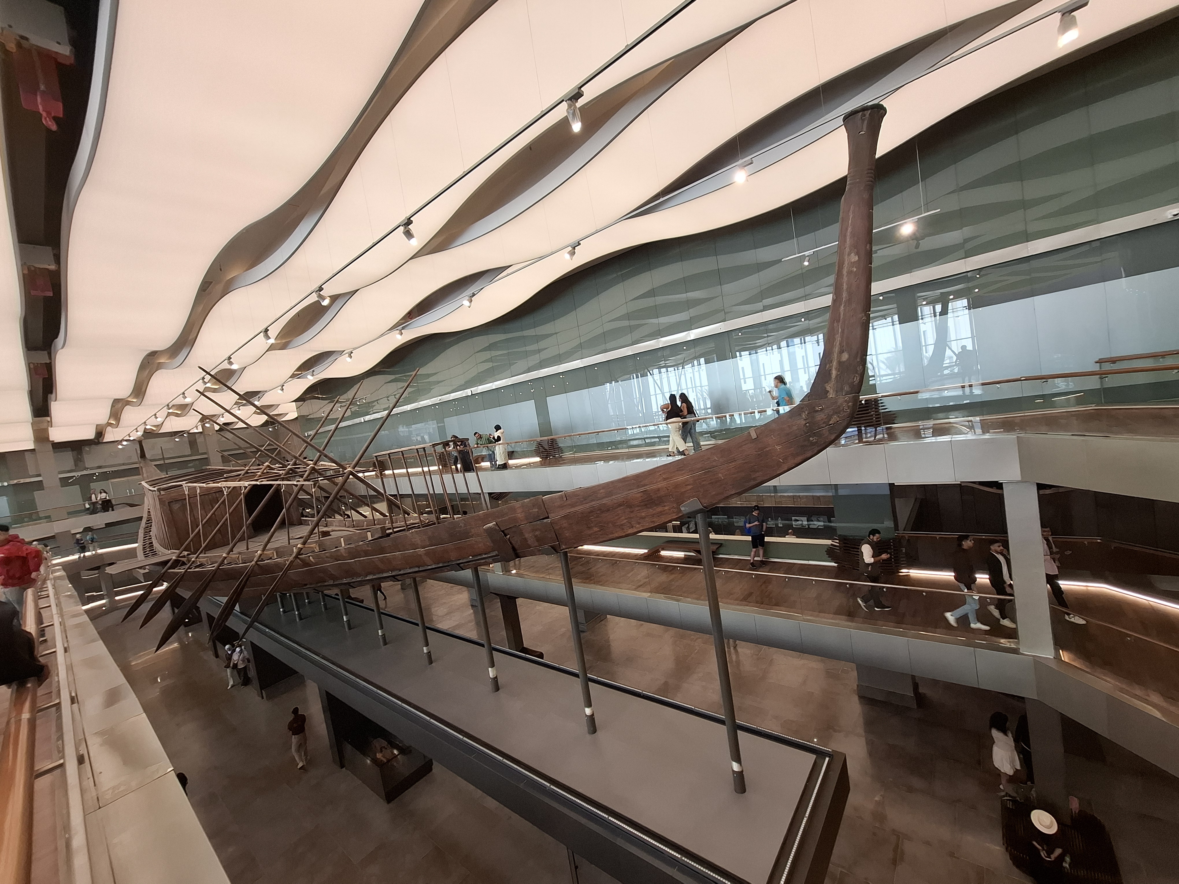 Interior view of the Grand Egyptian Museum showcasing the solar boat of King Khufu, with visitors observing the exhibit in a modern architectural setting.
