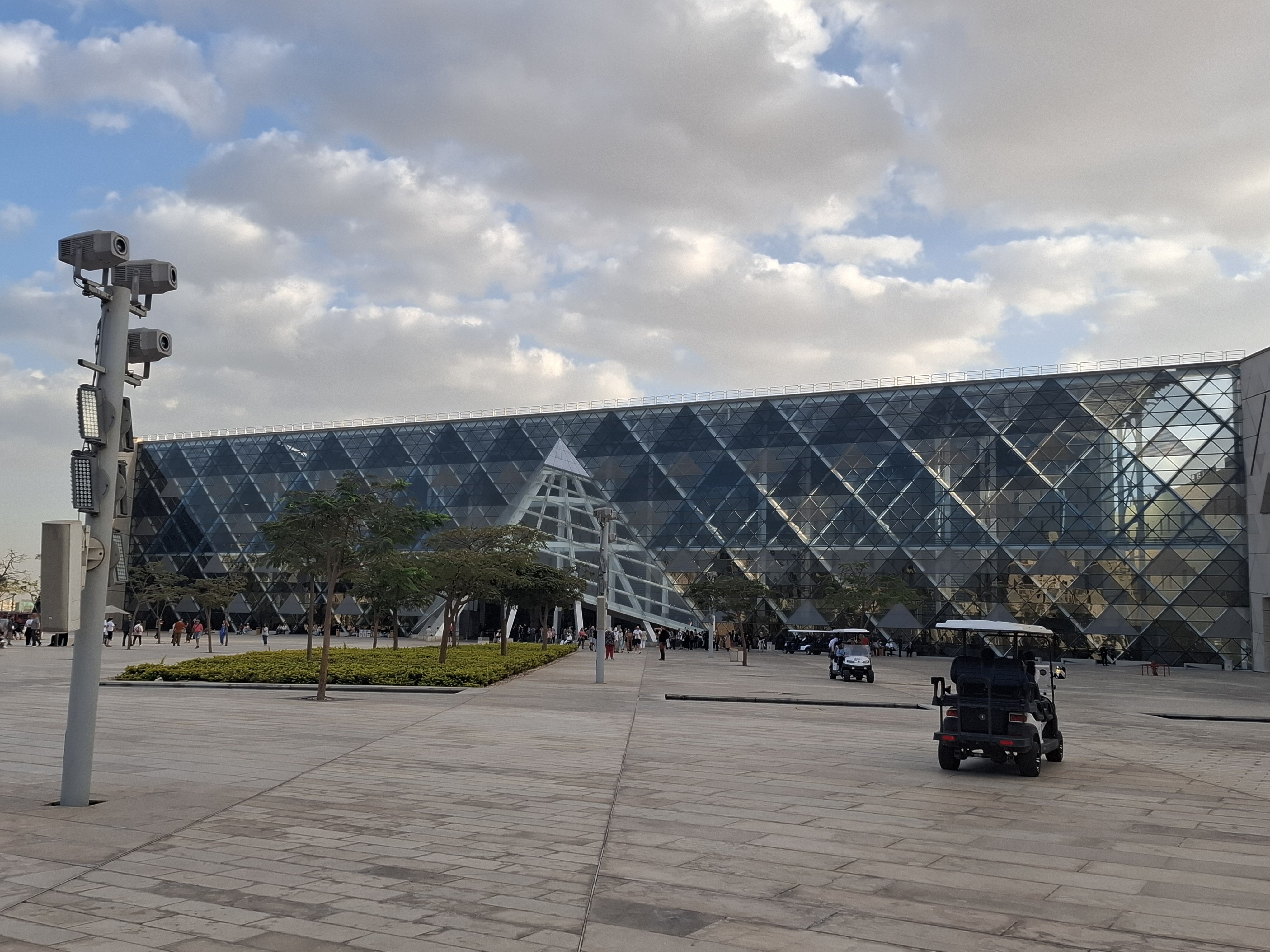 Exterior view of the Grand Egyptian Museum featuring a modern glass facade with a pyramid-shaped structure, surrounded by landscaped gardens and visitors.