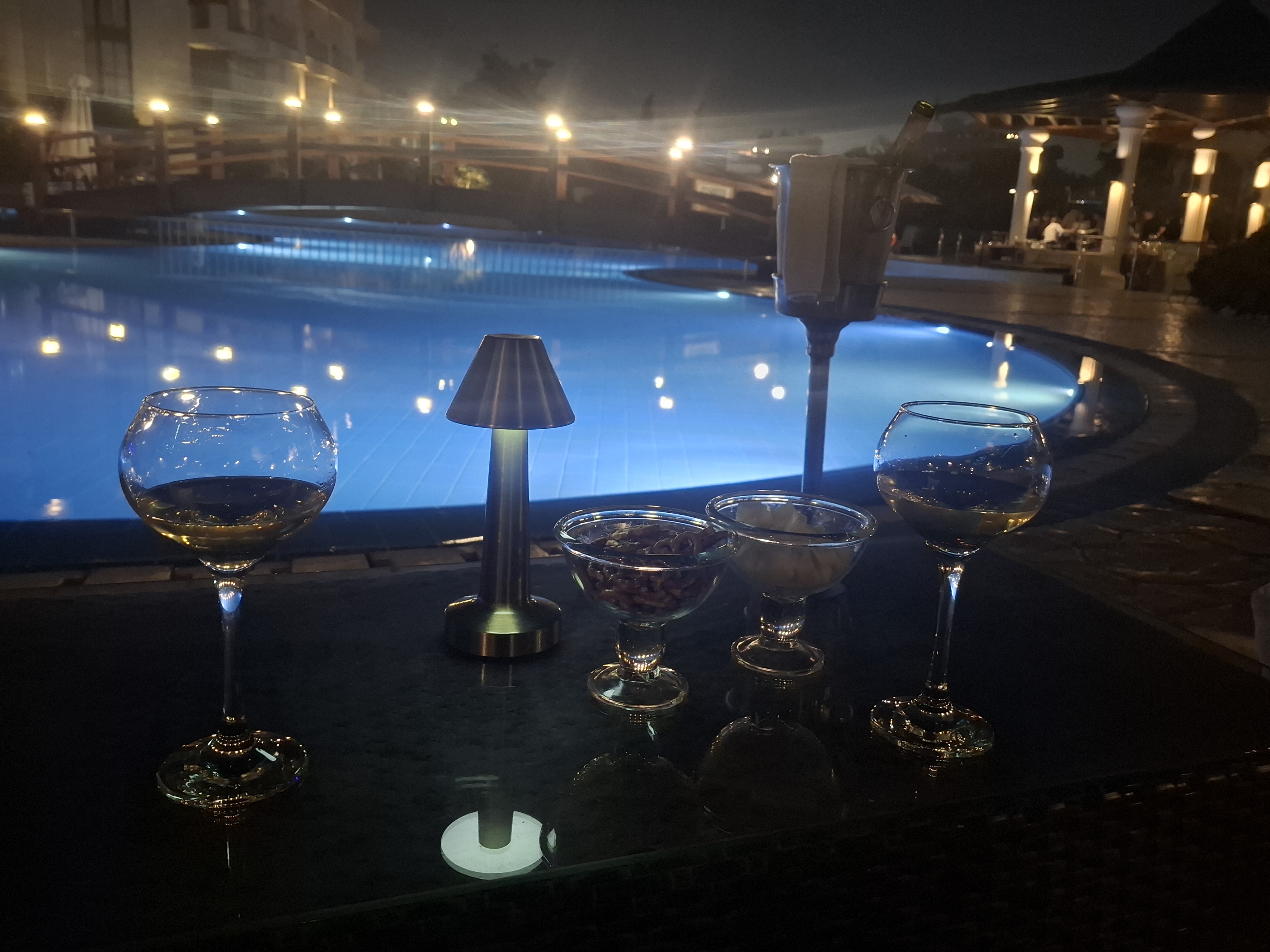 Two glasses of wine beside a small table with snacks, overlooking a lit swimming pool at night.