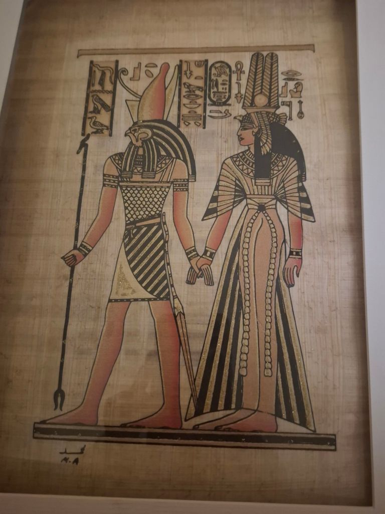 An illustration of two ancient Egyptian deities, featuring a male figure with a red crown and a female figure with a tall black crown, both adorned in traditional attire. They are depicted holding hands, surrounded by hieroglyphs on papyrus.