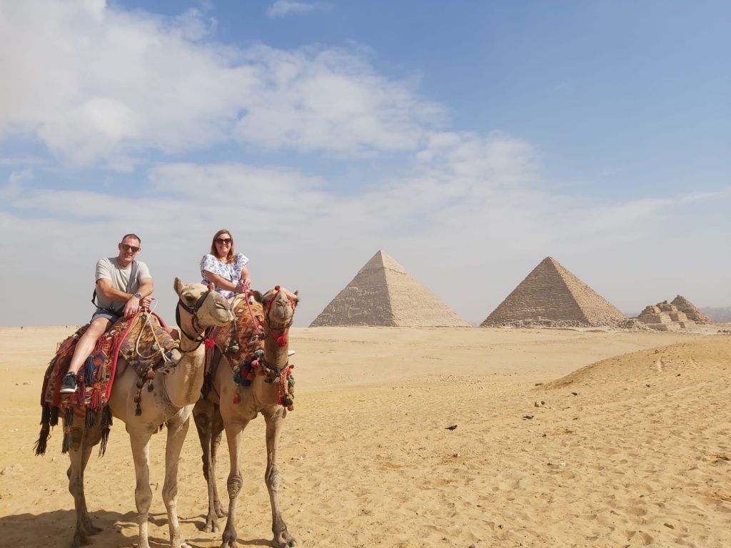 A couple riding camels in the desert near the Pyramids of Giza, with the pyramids visible in the background under a clear blue sky.