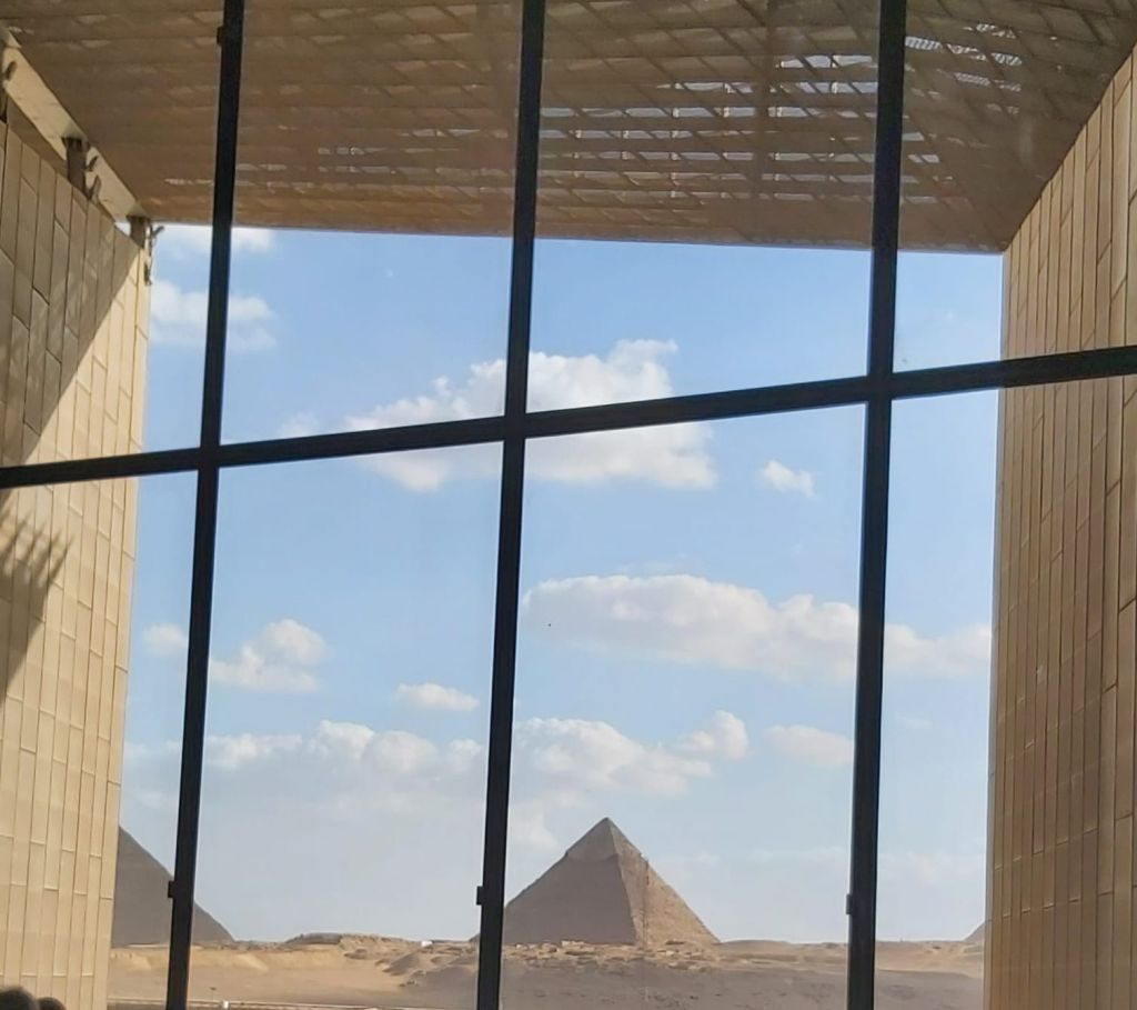 View of the Pyramids of Giza through a large glass window, with clouds in the sky and desert landscape visible.